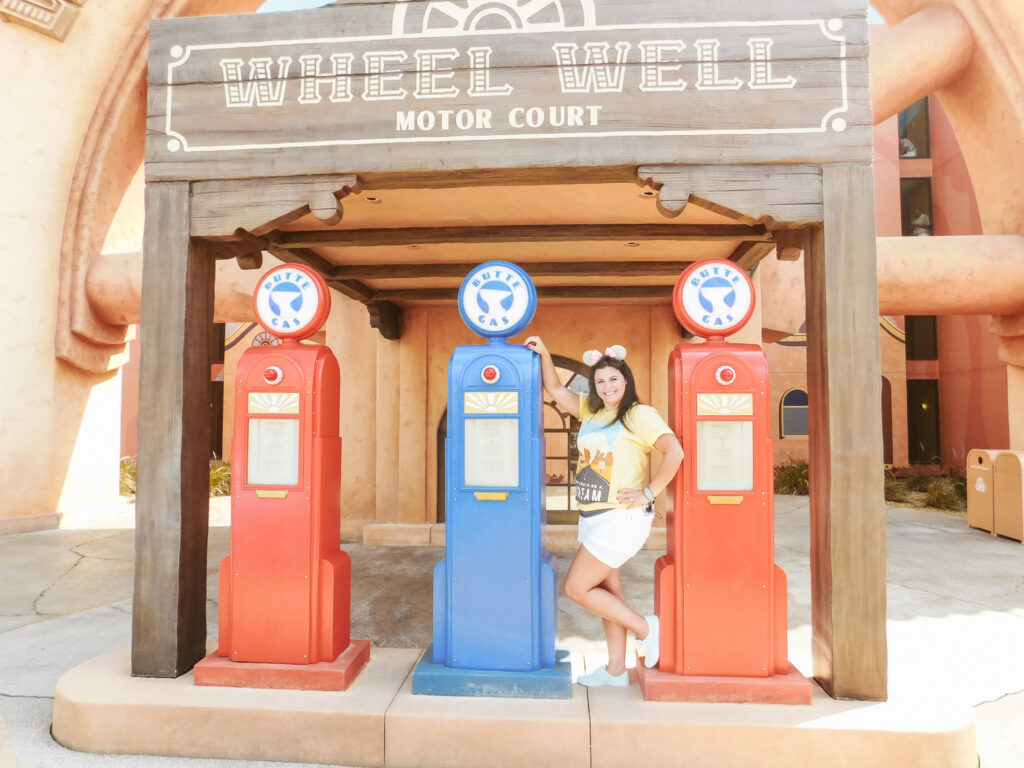 A young woman with brown hair and a Mickey ears headband posing next to old-fashioned gas pumps with a wooden sign above them that reads "Wheel Well Motor Court."