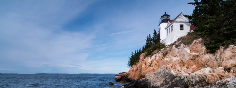A rocky shoreline with a small white building and lighthouse surrounded by trees.