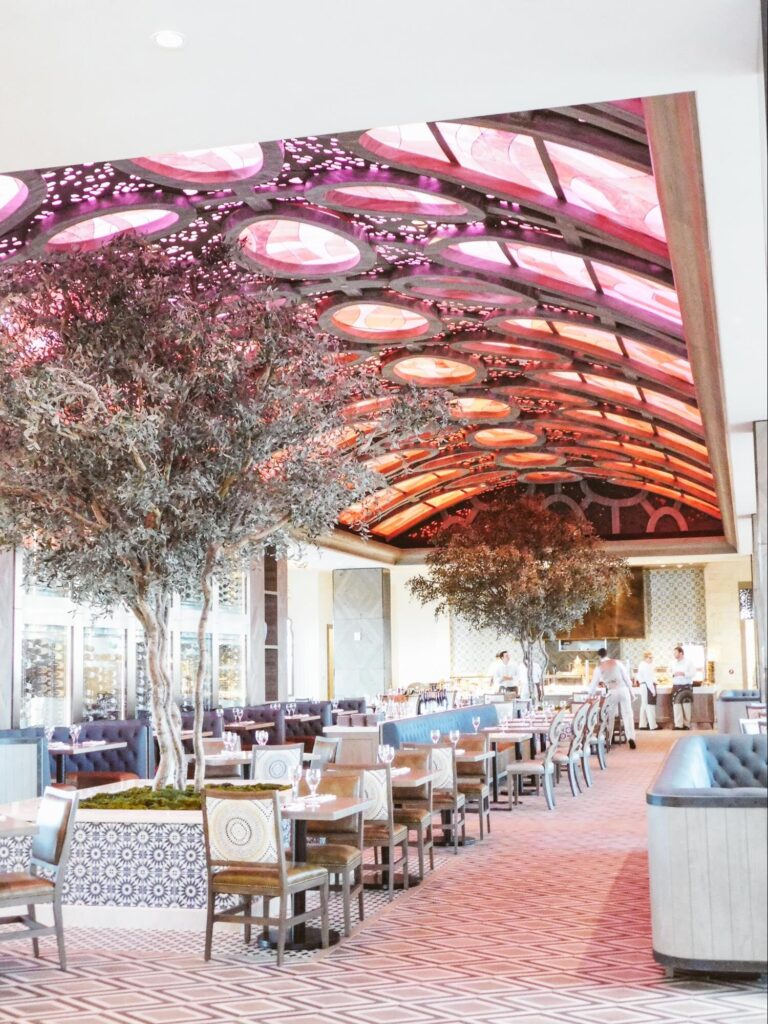 A large, bright, rectangular dining room with an arched glass ceiling and trees growing in two planters near the center.