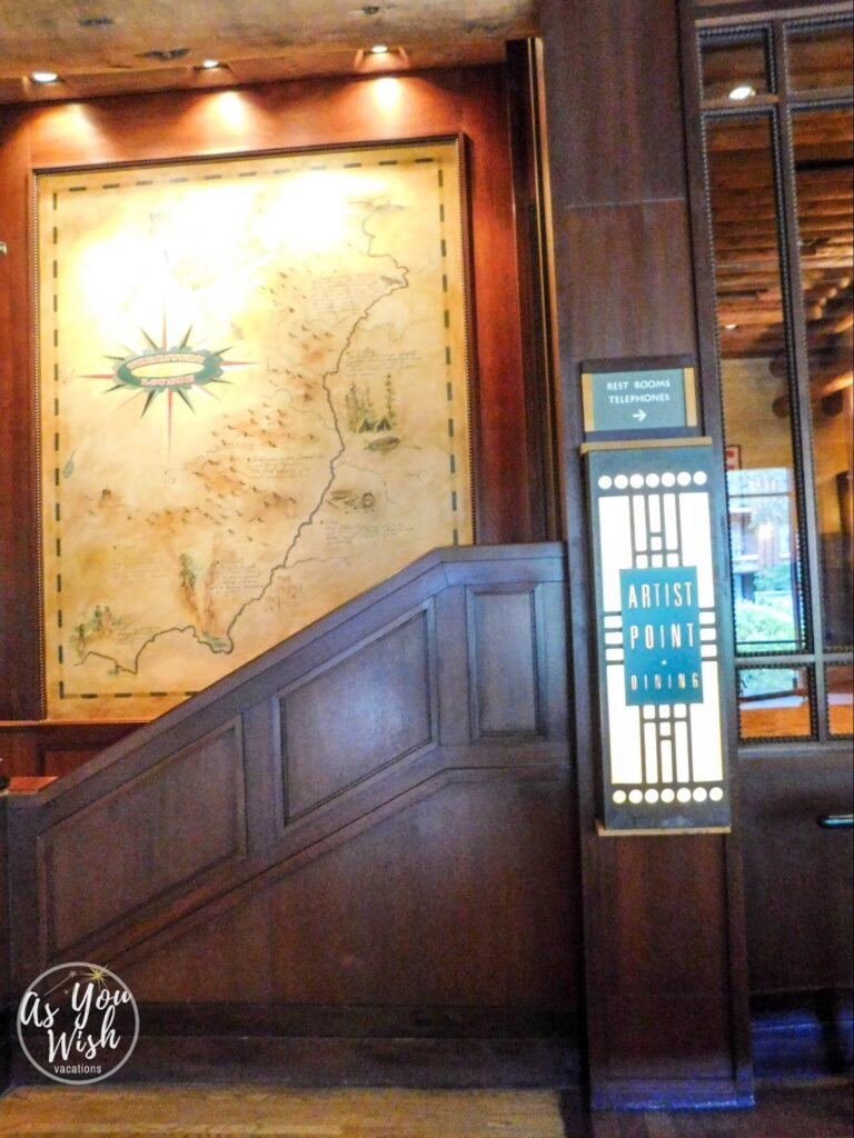 The side of a dark wood staircase with a hand-drawn map on the wall next to it. In front of the staircase is an art deco-style sign which reads "Artist Point Dining."