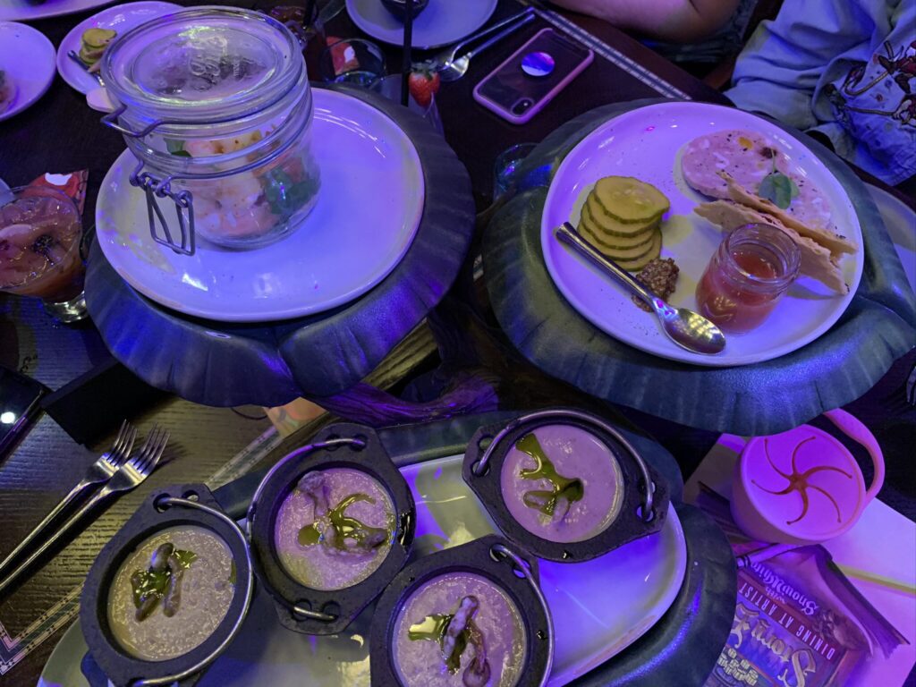 A meal served on serving trays made to look like lily pads. The meal features mushroom bisque, shrimp, lavosh bread, chicken terrine, pickles, and stone fruit preserves.