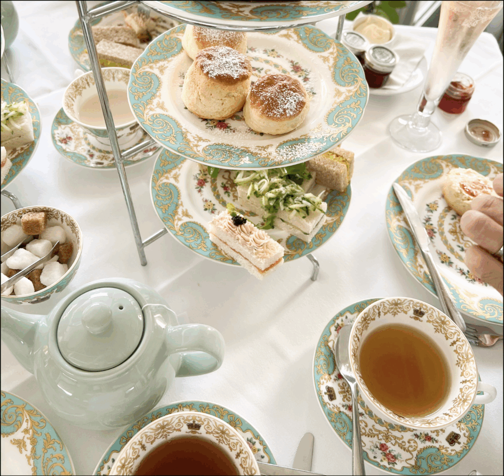A breakfast spread featuring American biscuits (or, in British English, scones), finger sandwiches, tea, and jam on a light blue tea set with blue and gold swirl and pink rose motifs.