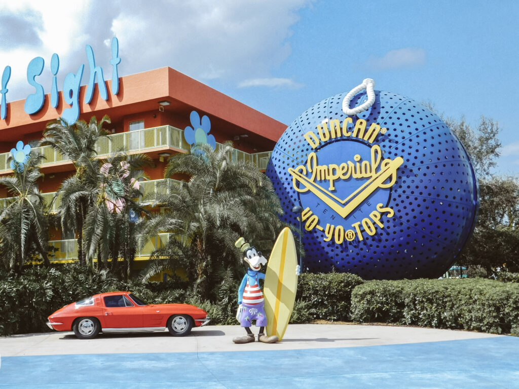 Disney mascot Goofy holding a surfboard in front of a vintage car and a giant yo-yo statue that reads "Duncan imperial yo-yo tops."
