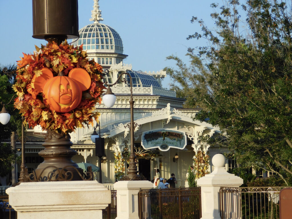 A wreath of fall leaves with a pumpkin shaped like Mickey Mouse in the center hangs on a lamp post in front of the Crystal Palace, an ornate white building with a domed glass roof.