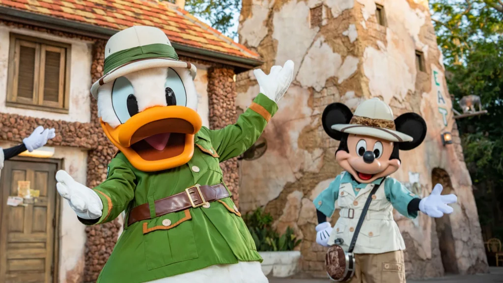 An area outside of Disney's Tusker House Restaurant. Donald Duck and Mickey, dressed in safari gear, gesture to the camera as if inviting the viewer over. In the background are two rustic-looking buildings made of beige stone.