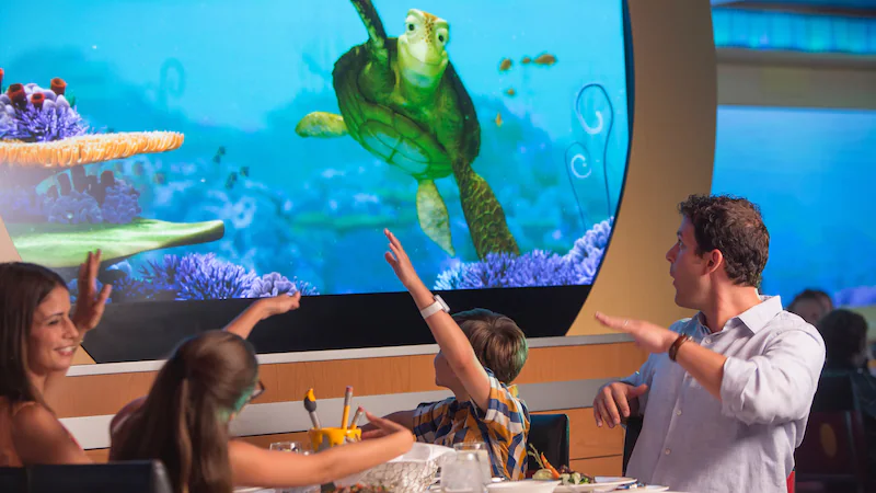 A family waving to a large photo or video screen with Crush, the turtle from Finding Nemo, during a meal.